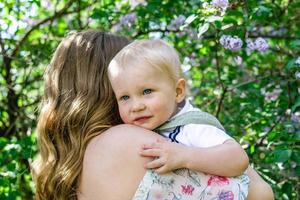 Portrait of mother with little son in her hands in park with lilac blossom tree, back view. Happy mother and child. photo