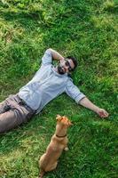 Man and dog lying on green grass in summer, top view. Attractive European man resting with his dog. photo