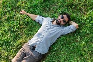 joven y guapo deportista europeo con gafas de sol descansa sobre un césped en el parque de verano, vista superior. foto