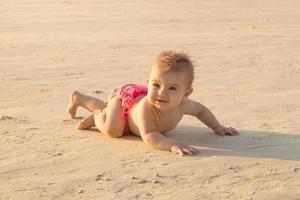 la pequeña niña se arrastra en una playa de arena cerca del mar a la luz del sol. foto