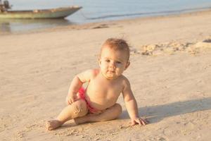 Cute little baby girl is sitting on a sandy beach near to sea in sunset sunlight. photo