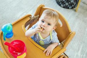 Adorable little baby girl is sitting in wooden baby chair after lunch and eating cookie. photo