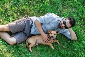 Attractive European man in casual clothing and sunglasses is lying on grass and hugging his cute little dog. photo