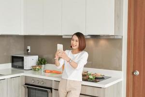 Young asian woman in the kitchen using mobile phone making video call while cooking meal photo