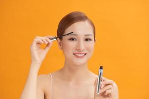 Beautiful young woman with eyelash extensions applying mascara, on yellow background photo