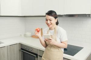 pretty young woman using her mobile phone while eating an apple in the kitchen at home. photo