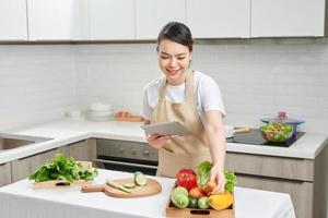 beautiful korean woman in apron holding tablet pad looking at list and checking fresh vegetables. photo