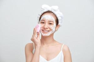 Happy young Asian woman applying foaming cleanser on her face and wearing towel on her head. photo