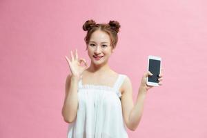 Attractive young girl showing ok gesture while holding blank screen mobile phone isolated over pink background photo