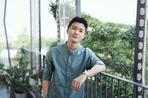 A portrait of a handsome young adult male that is leaning on a railing, with the sun shining and trees around. photo