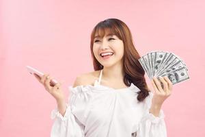 Photo of pleased happy screaming young woman posing isolated over pink wall background using mobile phone holding money.