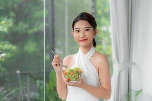 atractiva mujer sonriente asiática aislada en la sala de estar en casa comiendo ensalada foto