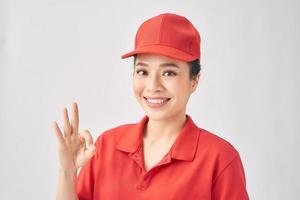 Portrait of attractive delivery woman in red t-shirt and cap smiling and showing okey sign isolated over pink background photo