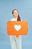 heerful young female in lilac and white clothes holding in hand red speech bubble with heart icon photo