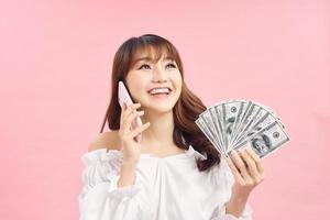 Happy Shocked ginger woman talking by smartphone while holding money and looking at the camera over pink background photo