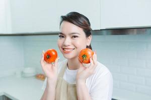 Cheery girl holding in hands tomato cooking everyday in modern light white interior kitchen photo