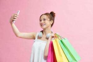 Excited asian shopaholic girl using phone posing with shopping bags on pink background. Sales and discounts season. photo