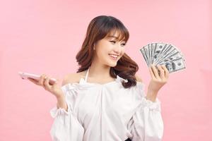 imagen de una hermosa joven emocionada posando aislada sobre un fondo de pared rosa sosteniendo dinero y usando un teléfono móvil. foto