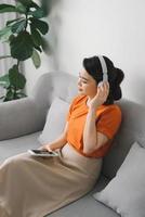 Side view of a smiling pretty woman in headphones using mobile phone while sitting on a sofa at home photo