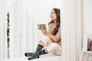 Happy beautiful woman drinking hot coffee sitting on window sill in christmas decorated home. Holiday concept photo