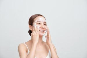 Happy attractive young Asian woman applying foaming cleanser on her face over white background. photo