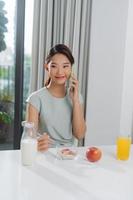 Beautiful woman eating tasty oat with milk while talking on mobile phone at table photo