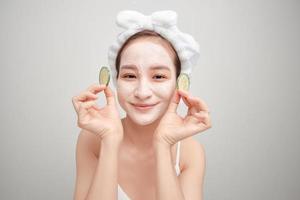 Beautiful young woman with facial mask on her face holding slices of fresh cucumber. photo