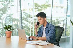 man talking on mobile phone, working from home. Portrait of pensive asian businessman using laptop computer, planning project, communication in modern office photo