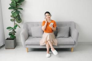 Side view of a smiling pretty woman in headphones using mobile phone while sitting on a sofa at home photo