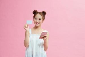 Beautiful happy young woman isolated over pink background, showing plastic credit card while using mobile phone photo