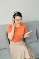 mujer atractiva asiática usando auriculares, usando un teléfono móvil y sentada en el sofá foto