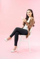 Portrait of a smiling beautiful girl wearing dress holding mobile phone while sitting on a chair isolated over pink background photo