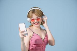 Photo of excited woman in swimwear isolated over blue wall background showing display of mobile phone listening music with headphones.