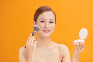 Beautiful woman applying blusher to her cheek with a large cosmetics brush while holding a compact mirror in front of her isolated on yellow photo