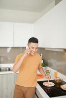Handsome guy is talking on the mobile phone and smiling while cooking in the kitchen photo