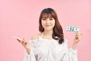 Portrait of a smiling young girl in dress standing over pink background, holding mobile phone, showing money banknotes photo