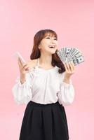 Portrait of a smiling young girl in dress standing over pink background, holding mobile phone, showing money banknotes photo