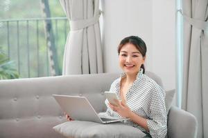 Photo of smiling pregnant asian woman using laptop and mobile phone while sitting on couch at home