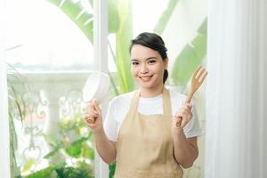 Woman in apron in hand frying pan and kitchen spatula near big window photo