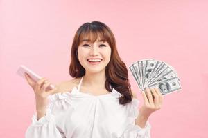 Portrait of an excited happy girl showing bunch of money banknotes while holding mobile phone and looking at camera isolated over pink background photo