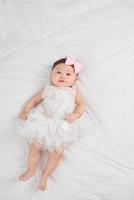 Niña pequeña sonriente con vestido blanco y decoración de lazos para el cabello acostada en la cama foto