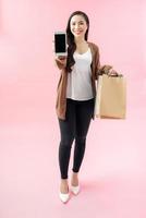 Full length portrait of a beautiful young woman standing isolated over blue background, carrying shopping bags, showing blak screen mobile phone photo