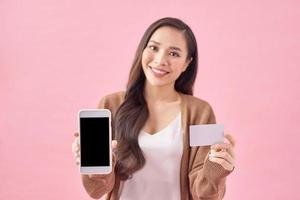 imagen de una joven asiática feliz parada aislada sobre un fondo rosado. mirando a la cámara mientras usa el teléfono y tiene una tarjeta de débito. foto