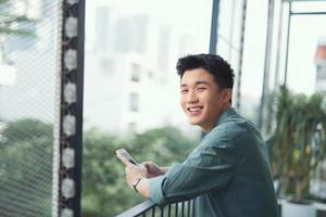 estudiante masculino asiático enviando mensajes de texto en un teléfono inteligente en el balcón foto