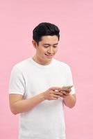 Waist-up portrait of joyful Vietnamese man leaning on gray wall while browsing Internet on smartphone, studio shot photo
