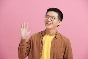 Asian happy young handsome man in casual clothing waving hand greeting say hello hi of goodbye on pink background. photo