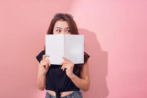 retrato de una linda joven escondida detrás de un libro abierto y mirando hacia otro lado aislada sobre el fondo de la pared rosa foto