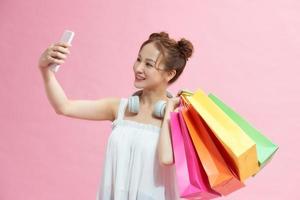 Happy young woman with shopping bags taking selfie on pink background photo