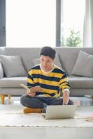hombre asiático sentado en el suelo y haciendo cálculos financieros negocios en línea en un nuevo apartamento foto