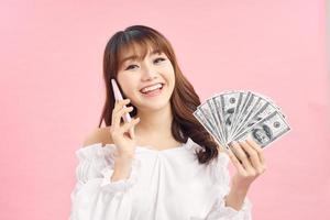 Portrait of a cheerful young woman holding money banknotes and celebrating isolated over pink background. Using mobile phone. photo